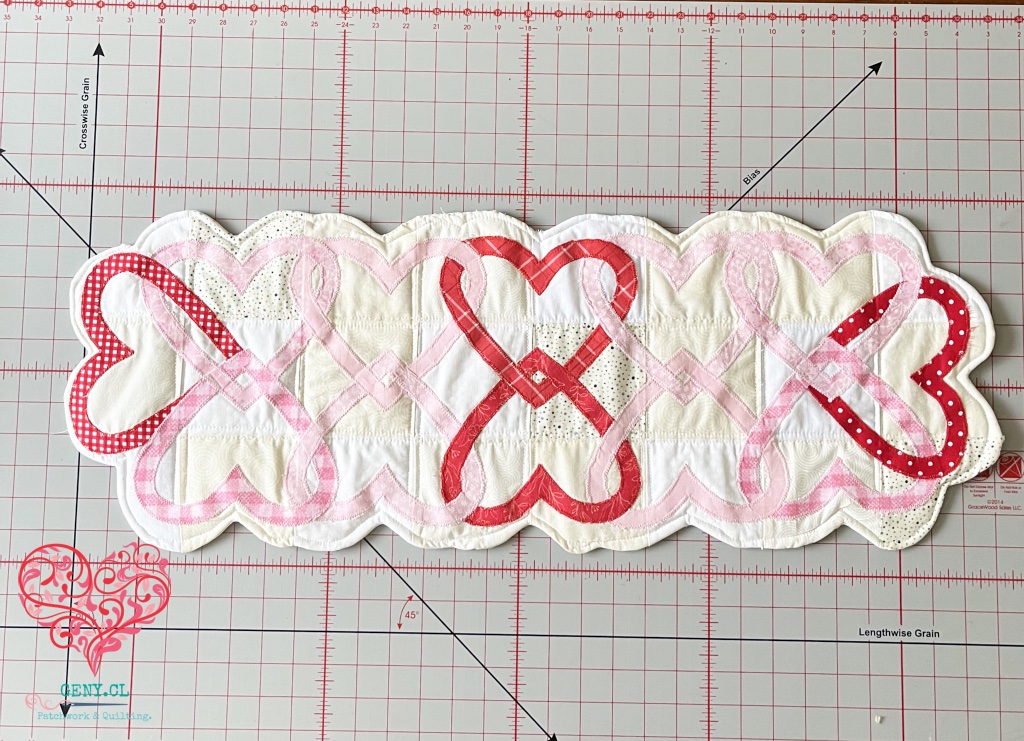 Hearts Entwined Table Runner de Art to Heart terminado, con corazones entrelazados en rojo y rosa y borde curvo acolchado.