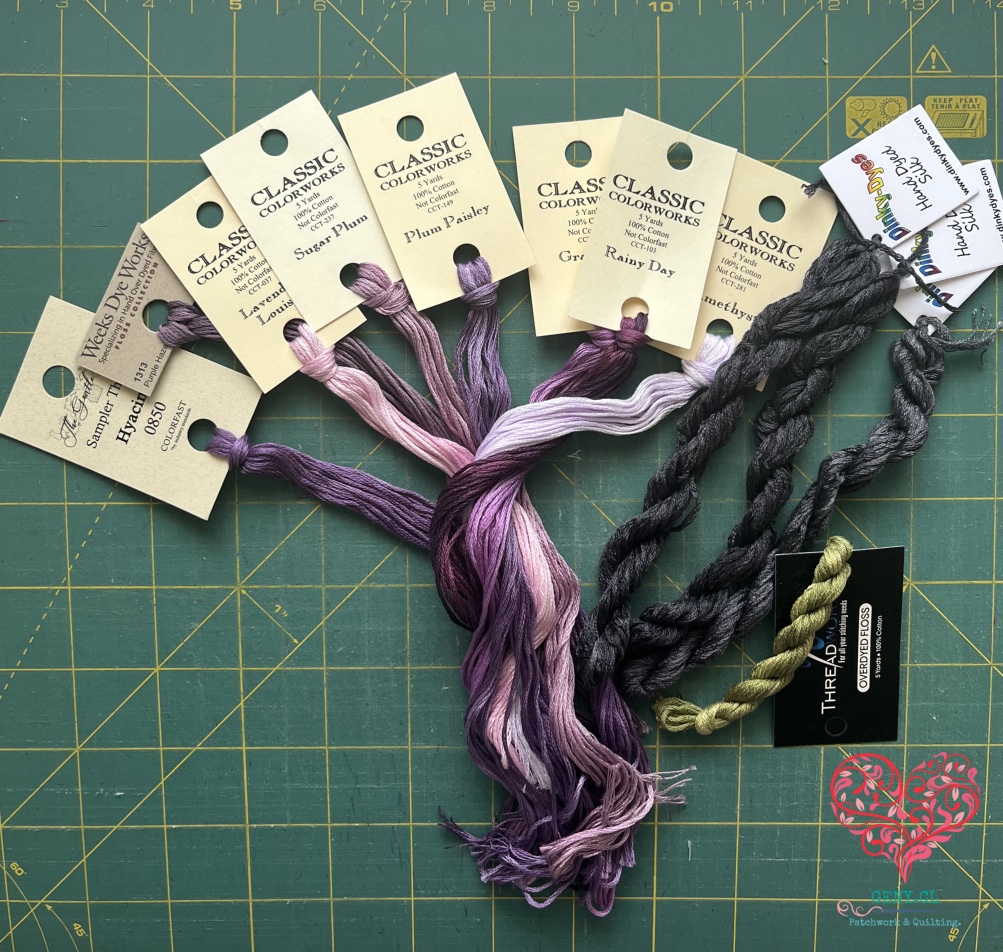 Madejas de hilos teñidos a mano en tonos morados, lilas, rosados, grises y verde oliva, de varias marcas, dispuestas sobre un mat de corte verde.