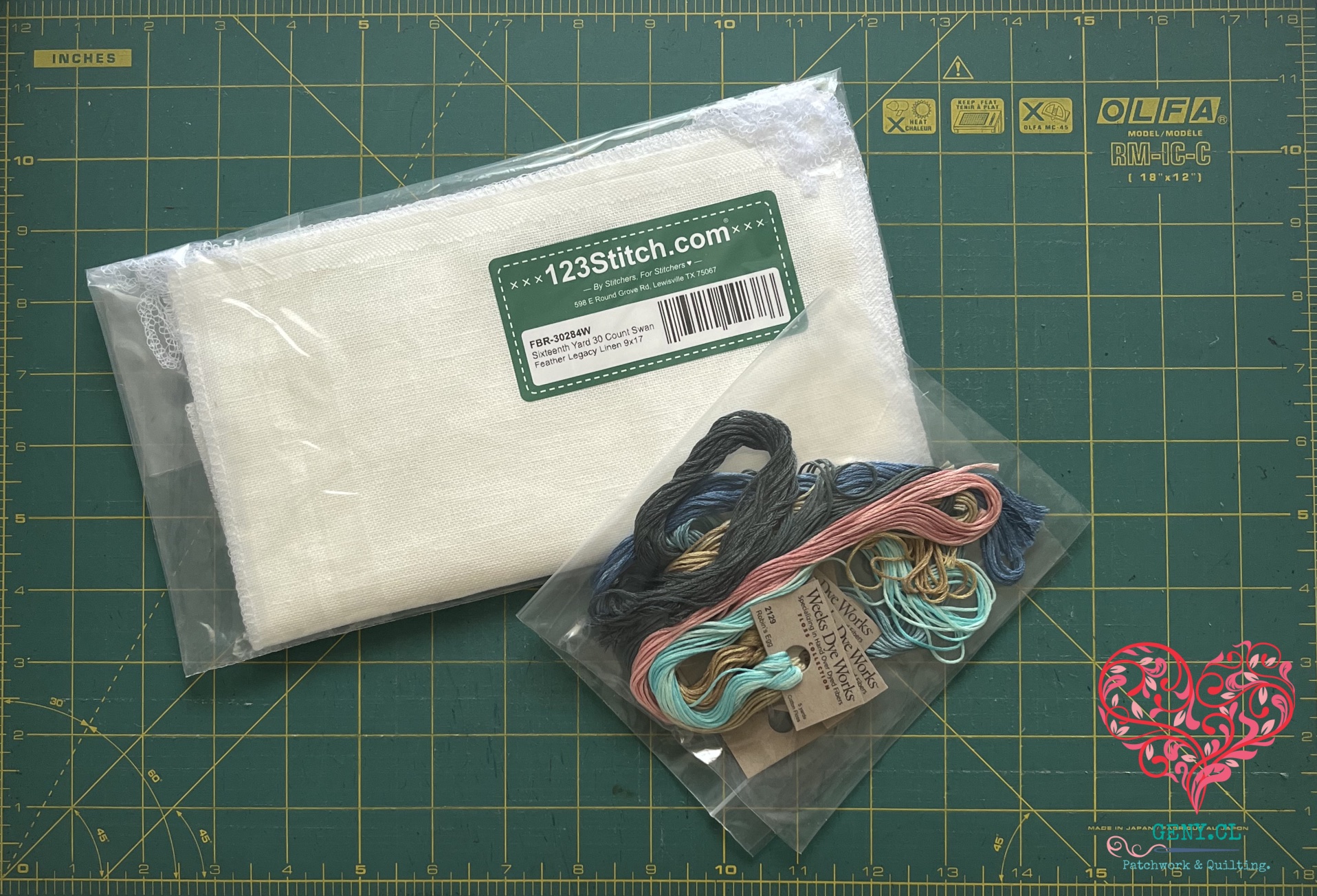 Materiales para un futuro bordado de punto de cruz: tela Feather Legacy Linen color Swan e hilos de bordado en tonos azules, rosados, turquesas y oscuros sobre un mat de corte verde.
